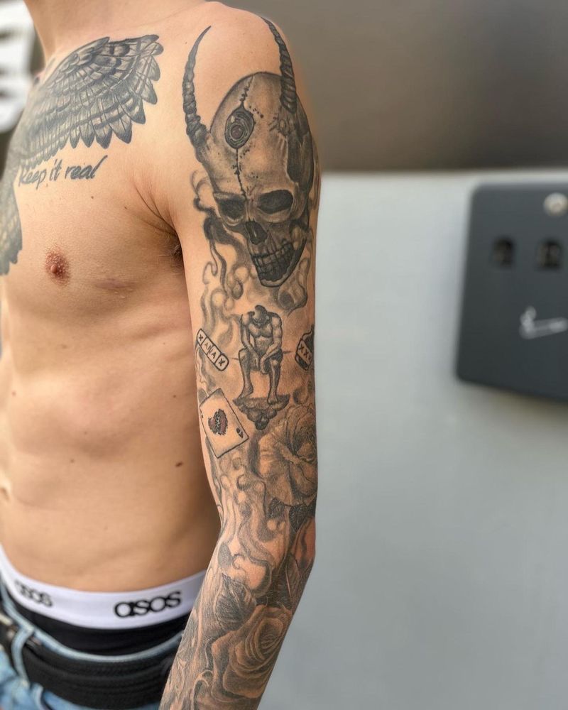 Black and grey full sleeve: a horned skull over smoke, a thinker figure, cards and roses